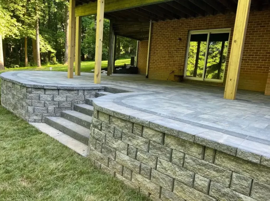 A newly constructed outdoor patio with stone steps and retaining walls, covered by a wooden structure, attached to a brick house in a grassy backyard.