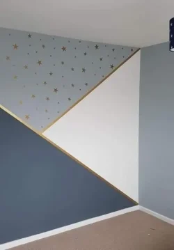 A room corner featuring a wall with blue, white, and dark grey triangular sections and gold star decals, separated by gold lines. The floor is carpeted in light brown, and the ceiling is white.