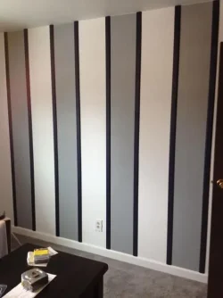 A wall with alternating vertical stripes of light gray, white, and dark gray colors, with an electrical outlet near the bottom. A dark table with items on it is partially visible in the foreground.