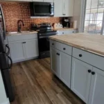 A modern kitchen with white cabinets, black appliances, a wood countertop island, and a copper backsplash. The floor is wood, and a ceiling fan with lights is above. A large window brings in natural light.