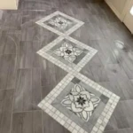 A tiled floor with grey square tiles arranged diagonally, featuring three large floral-patterned tiles centrally aligned, and a brick wall at the back.