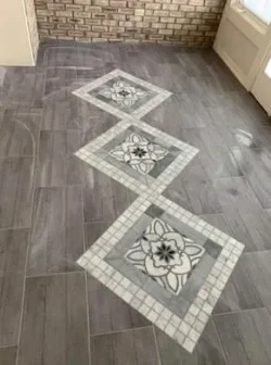 A tiled floor with grey square tiles arranged diagonally, featuring three large floral-patterned tiles centrally aligned, and a brick wall at the back.