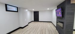 A basement room with light wood flooring, white walls, a small window, a black door, and a wall-mounted TV next to a fireplace.
