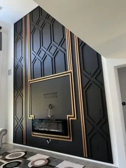 A modern black accent wall with geometric patterns and gold trim features a built-in electric fireplace and TV mount, located in a room with a patterned rug and white walls.