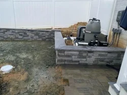 A partially constructed swimming pool with an unfinished concrete area, paving stones, and pool equipment installed near the edge, all enclosed by a white privacy fence.