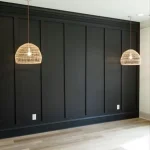 An empty room with a black, paneled wall, two hanging rattan pendant lights, light wood flooring, and two electrical outlets on the wall.