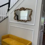 A yellow bench is placed against a white wall beneath a decorative mirror. The wall leads to a staircase with white balusters and a dark handrail.