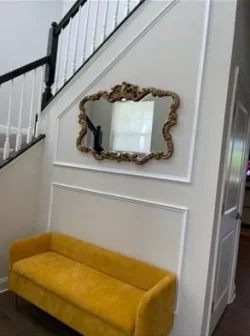 A yellow bench is placed against a white wall beneath a decorative mirror. The wall leads to a staircase with white balusters and a dark handrail.